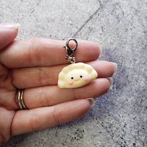 May include: A small, white, smiling dumpling charm with a gold clasp. The charm is made of polymer clay and has a cute, cartoon-like face.