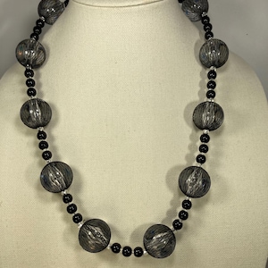 May include: A black and silver necklace with large, round, black mesh beads. The necklace is displayed on a white mannequin.
