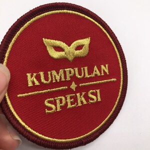 Puede incluir: Un parche circular bordado de color rojo con una máscara dorada, las palabras "KUMPULAN" y "SPEKSI" en hilo dorado.