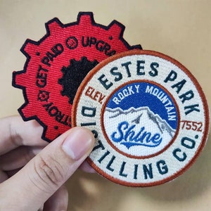 May include: Two embroidered patches. One is red with black text that reads "Get Paid" and "Upgraded" in a gear shape. The other is a round beige patch with a blue mountain scene and the text "Estes Park Rocky Mountain Dist. Co. Elev. 7552" and "Shine" in a white circle.