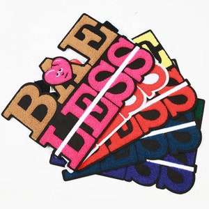May include: A stack of colourful embroidered patches with the words "BE", "I LOVE", "SO", "GOOD", "YOU", and "DAD" in various colours. The patches are made of a soft, fuzzy material and have a white border.