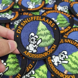 May include: A close-up of a blue and orange embroidered patch featuring a white bunny rabbit holding a branch of a green pine tree. The text on the patch reads "De Snuf fellaars Abdissenbosch".