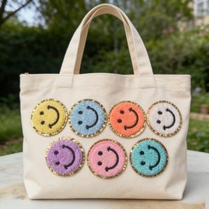 Peut inclure: Sac fourre-tout en toile de couleur crème orné de huit écussons smiley colorés. Chaque smiley est de couleur différente : jaune, bleu, orange, blanc, violet, rose et turquoise, avec une bordure dorée. Le sac a deux anses et une fermeture éclair.