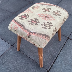 May include: A small, wooden stool with a patterned cushion. The cushion is made of a woven fabric with pink, brown, and white geometric designs. The stool has a simple, rustic look.