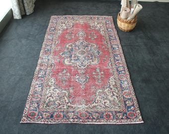 Puede incluir: Una alfombra rectangular de aspecto envejecido, con un campo central rojo y complejos diseños florales azules y crema. La alfombra tiene un borde azul oscuro con motivos florales y probablemente es de lana. Se encuentra sobre una superficie gris oscuro.