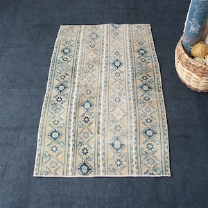 Può includere: Tappeto rettangolare beige con strisce verticali di motivi geometrici blu. Il tappeto ha un aspetto invecchiato. Un cestino di vimini con filato e tessuti arrotolati è nelle vicinanze.