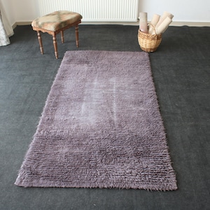 May include: A rectangular, shaggy, mauve-colored rug with a textured surface. The rug is placed on a dark gray floor. A small wooden stool with a patterned seat and a wicker basket with rolled-up textiles are also visible.