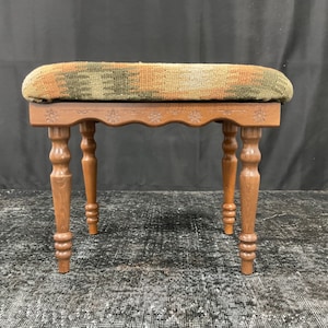 May include: A wooden stool with a woven fabric seat in shades of green, brown, and orange. The stool has four turned legs with decorative details.