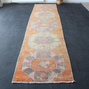 May include: An orange and gray patterned runner rug with geometric designs. The rug features a central pattern of repeating shapes in shades of orange, gray, and pink. The rug is rectangular and appears to be made of woven material.