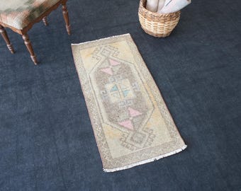 Puede incluir: Una alfombra rectangular de estilo vintage con un diseño geométrico central en marrón, beige y rosa pastel y azul. La alfombra tiene un borde con flecos y está colocada sobre una superficie oscura. Una pequeña silla de madera y una cesta de mimbre están en el fondo.