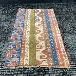 Op de afbeelding: Een lang, smal tapijt met een kleurrijk geometrisch patroon. Het tapijt heeft aan beide uiteinden franjes en is gemaakt van wol. De kleuren in het tapijt zijn rood, blauw, groen en wit.