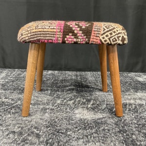 May include: A small, wooden stool with four legs. The seat is covered in a colourful, patterned fabric with shades of brown, pink, and white.