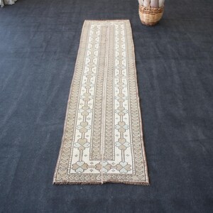 May include: A rectangular, beige and cream-colored runner rug with a repeating floral and geometric pattern. The rug has a central design with vertical stripes and a border. The rug is approximately 2 feet wide and 6 feet long.