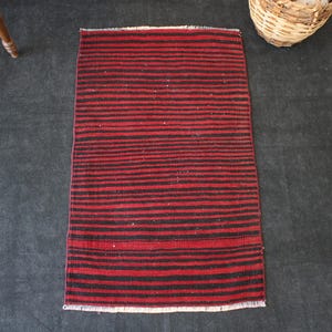 May include: A rectangular woven rug featuring a repeating pattern of horizontal stripes in deep red and black. The rug has a flat-weave texture and includes short, light-coloured fringe detailing along the top and bottom edges of the piece.
