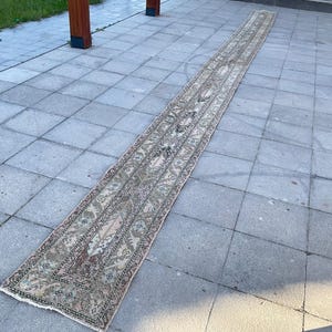 Puede incluir: Una alfombra larga y estrecha con un intrincado diseño floral en tonos beige, rosa y verde. La alfombra está colocada sobre una superficie de baldosas grises, mostrando su longitud y su diseño detallado. Los bordes de la alfombra están ligeramente deshilachados.