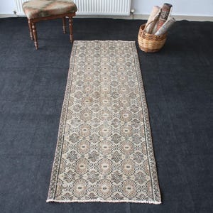 May include: A rectangular runner rug featuring a repeating geometric pattern in shades of beige, brown, and charcoal gray. The rug has a low-pile texture with a symmetrical design and short fringe detailing along the two shorter ends of the piece.