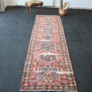 May include: A long, rectangular runner rug featuring a faded red base color with intricate geometric patterns in shades of blue, yellow, and dark brown. The design includes repeating central medallions and a detailed border with small motifs.