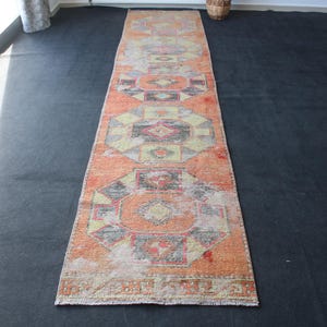 May include: An orange and beige runner rug with geometric patterns. The rug features a central design of octagons and diamonds in shades of red, black, and yellow. The rug is rectangular and appears to be made of woven material.