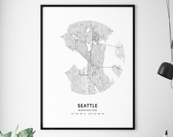 May include: A black and white print of a map of Seattle, Washington, in a black frame hanging on a white wall. The map is in a circle and includes the city's streets and landmarks. The print is hanging above a gray desk with two gray chairs and two black computer monitors.