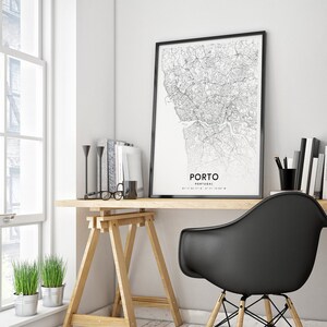 May include: A black and white map print of Porto, Portugal, framed in a black frame, hangs above a wooden desk with a black chair. The desk has a wooden easel and a pair of glasses on top.