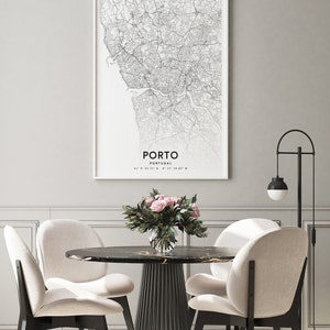 May include: A black and white map print of Porto, Portugal, with the city name and coordinates printed below the map. The print is framed and hanging on a gray wall above a dining table with four chairs. A vase of pink roses sits on the table.