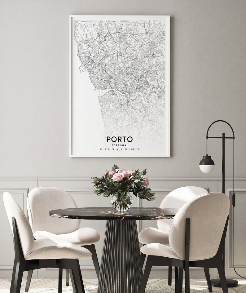 May include: A black and white map print of Porto, Portugal, with the city name and coordinates printed below the map. The print is framed and hanging on a gray wall above a dining table with four chairs. A vase of pink roses sits on the table.