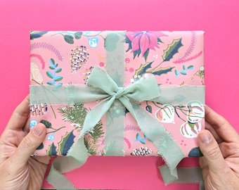 Könnte beinhalten: Ein rechteckiges Geschenkpaket, eingewickelt in rosafarbenes Papier mit einem botanischen Muster aus Tannenzapfen, Stechpalmenblättern und Blumen. Das Paket ist mit einem salbeigrünen, ausgefransten Band zu einer Schleife gebunden.