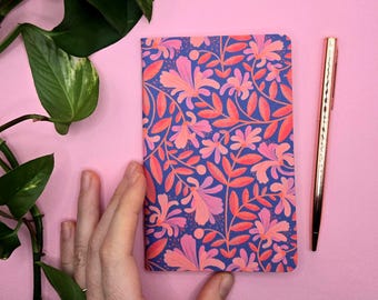 Könnte beinhalten: Ein Notizbuch mit floralem Muster mit blauem Hintergrund und rosa und orangefarbenen Blumen. Ein goldener Stift liegt neben dem Notizbuch auf einer rosa Oberfläche. Grüne Blätter befinden sich in der oberen linken Ecke.