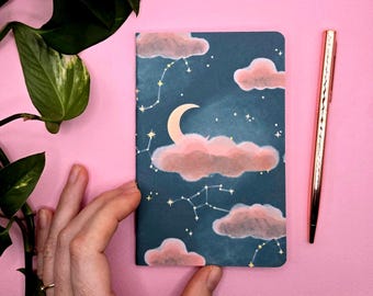 Könnte beinhalten: Ein kleines Notizbuch mit einem Himmelsdesign mit einem Mond, rosa Wolken und Sternbildern auf dunkelblauem Hintergrund. Ein roségoldener Stift liegt neben dem Notizbuch auf einer rosa Oberfläche.