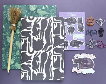 Könnte beinhalten: Ein Schreibwarenset bestehend aus einem dunkelgrün gemusterten Notizbuch, einem weißen Notizbuch mit schwarzen Katzensilhouetten, einem hellvioletten Stickerbogen mit Katzenmotiven und dem Text No tricks Just treats, sechs Büroklammern und einem Katzensticker.