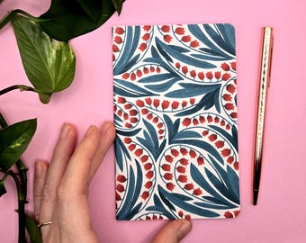 Könnte beinhalten: Ein Notizbuch mit floralem Muster, weißem Hintergrund, blaugrünen Blättern und roten glockenförmigen Blüten. Ein goldener Stift liegt neben dem Notizbuch auf einer rosa Oberfläche. Eine Hand hält das Notizbuch.
