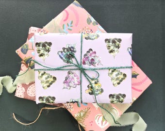 Könnte beinhalten: Ein rechteckiges Geschenk, eingewickelt in hellviolettes Papier mit einem wiederkehrenden Muster aus aquarellartigen Bäumen in Grün- und Violetttönen, gebunden mit dünner dunkelgrüner Schnur. Es liegt auf zwei weiteren rosa verpackten Geschenken.
