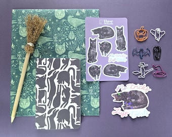 Könnte beinhalten: Ein Schreibwarenset bestehend aus grün gemustertem Papier, einem Notizbuch mit schwarz-weißem Katzenmuster, einem Miniatur-Besenstift, einem lavendelfarbenen Aufkleberbogen mit schwarzen Katzen und verschiedenen geformten Metall-Büroklammern.