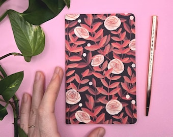 Könnte beinhalten: Ein Notizbuch mit floralem Muster mit roten und weißen Rosen auf schwarzem Hintergrund. Ein roségoldener Stift liegt neben dem Notizbuch auf einer rosa Oberfläche. Grüne Blätter sind in der oberen linken Ecke zu sehen.