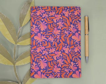 Könnte beinhalten: Ein rechteckiges Notizbuch mit einem lebendigen Blumenmuster aus rosa und orangefarbenen Blüten und Blättern auf dunkelviolettem Grund, begleitet von einem hellen Holzkugelschreiber, der rechts neben dem Notizbuch platziert ist.