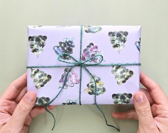 Könnte beinhalten: Ein rechteckiges Geschenk, eingewickelt in hellviolettes Papier mit einem sich wiederholenden Muster aus dunkelgrünen und violetten wolkenartigen Formen im Aquarellstil, gebunden mit einer dünnen dunkelgrünen Schnur.