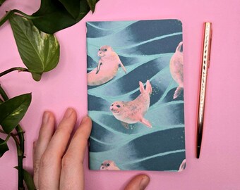 Könnte beinhalten: Ein rechteckiges Notizbuch mit einem sich wiederholenden Muster aus hellrosa Robben, die in dunkelblaugrünem Wasser schwimmen. Der Einband hat eine glatte Oberfläche und liegt neben einem metallischen roségoldenen Stift auf einer einfarbig rosa Fläche.