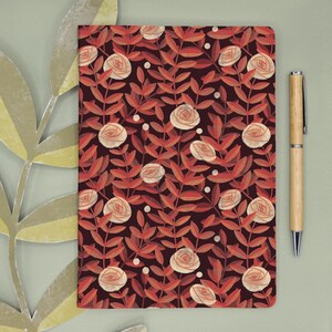Può includere: Un taccuino rettangolare con sfondo marrone scuro e un motivo ripetuto di rose color pesca chiaro e foglie marrone rossiccio, accompagnato da una penna a sfera in legno chiaro posizionata verticalmente a destra del taccuino.