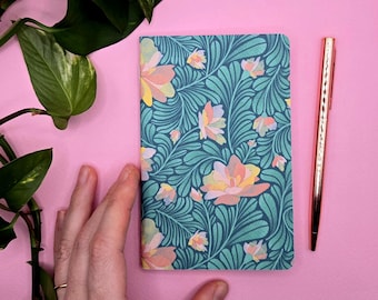 Könnte beinhalten: Ein Notizbuch mit türkisfarbenem Blumenmuster und einem roségoldenen Stift auf rosa Hintergrund. Das Notizbuch hat ein sich wiederholendes Design aus Blumen in Rosa-, Gelb- und Pfirsichtönen. Der Stift befindet sich vertikal neben dem Notizbuch.
