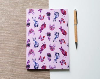 Könnte beinhalten: Ein Notizbuch mit rosa Einband, verziert mit Illustrationen von Katzen in Lila, Rosa und Blau. Ein Holzstift mit silberner Spitze liegt auf einer weißen Oberfläche neben dem Notizbuch.