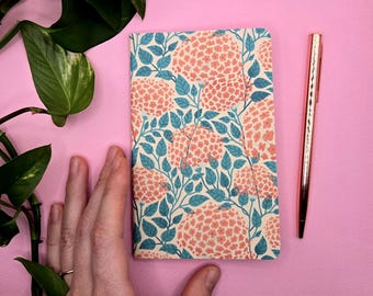 Könnte beinhalten: Ein Notizbuch mit floralem Muster und ein roségoldener Stift auf rosa Hintergrund. Das Notizbuch hat eine cremefarbene Basis mit korallenfarbenen Blumenclustern und türkisfarbenen Blättern. Der Stift befindet sich vertikal neben dem Notizbuch.