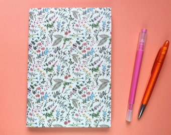 Könnte beinhalten: Ein Notizbuch mit floralem Muster auf weißem Hintergrund, mit einer Vielzahl bunter Blumen und grüner Blätter. Ein pinkfarbener Stift und ein orangefarbener Stift liegen auf einer korallenfarbenen Oberfläche neben dem Notizbuch. Schreibwaren.