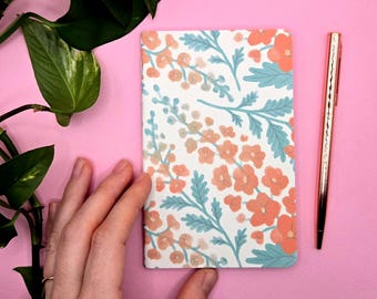 Könnte beinhalten: Ein Notizbuch mit floralem Muster in Orange und Blau, ein goldener Stift und eine grüne Pflanze auf rosa Hintergrund. Das Notizbuch hat einen weißen Hintergrund mit orangefarbenen Blumen und blauen Blättern.