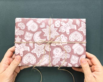 Könnte beinhalten: Ein rechteckiges Geschenk, eingewickelt in altrosa Papier mit einem sich wiederholenden weißen Muster aus Lebkuchenmännern, Schneeflocken, Fäustlingen, Herzen und Rentieren, befestigt mit einer dünnen braunen Schnur.