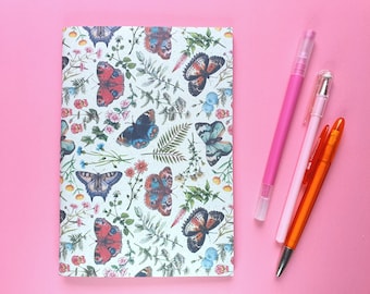 Könnte beinhalten: Ein Notizbuch mit einem Schmetterlings- und Blumenmuster, das Schmetterlinge in Blau, Orange und Braun sowie grüne Blätter und Blumen zeigt. Drei Stifte in Rosa und Orange liegen neben dem Notizbuch auf einem rosa Hintergrund.