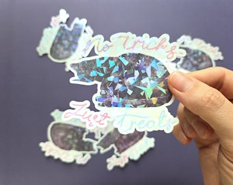 Könnte beinhalten: Ein holografischer Aufkleber in Form einer zusammengerollten Katze mit einem dunklen, reflektierenden, geometrischen Muster. Der Aufkleber zeigt den Text 'no tricks' über der Katze und 'just treats' darunter in einer hellen, kursiven, irisierenden Schrift.