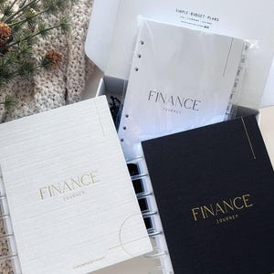 May include: Two spiral bound notebooks with gold lettering that reads "FINANCE JOURNEY" on the front cover. One notebook has a white cover with a subtle textured pattern and the other has a black cover. Both notebooks have a clear plastic cover with a tabbed divider.
