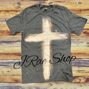 May include: A heather grey t-shirt with a white painted cross design. The cross is a simple design with a distressed look. The shirt has a crew neck and short sleeves. The text "J Rae Shop" is visible on the bottom of the shirt.