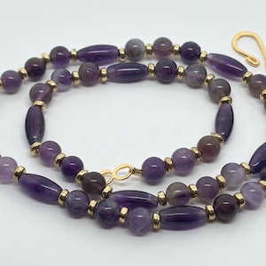 May include: A necklace featuring alternating purple amethyst beads in round and elongated oval shapes, separated by small gold-toned metal spacers. The piece is finished with a gold-toned hook-and-eye clasp mechanism at the ends of the strand.