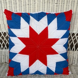 May include: A square quilted throw pillow featuring a central eight-pointed star pattern. The design is composed of red, white, and blue fabric segments stitched together in a geometric layout, creating a vibrant, multi-colored star motif.